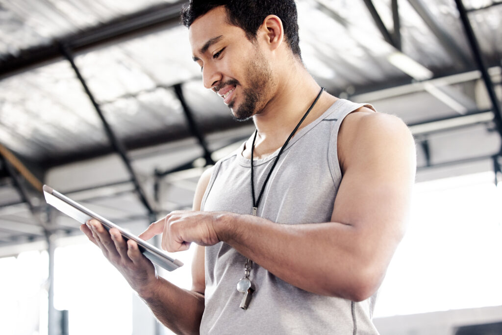 Male fitness trainer using a tablet to track client progress in a bright gym, wearing a whistle and casual workout attire.