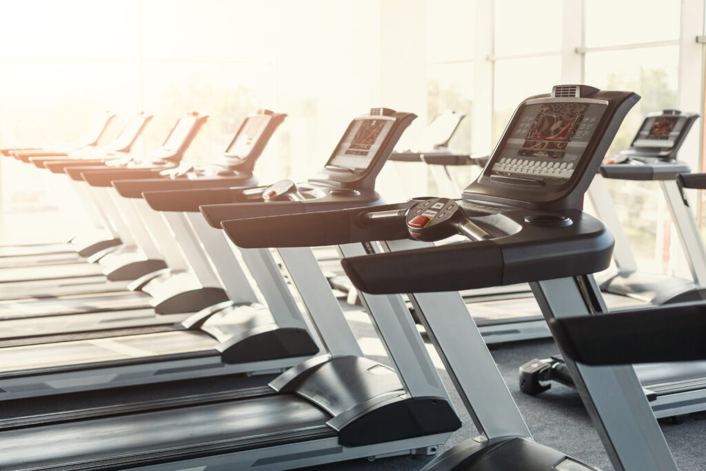 Row of modern treadmills in a well-lit gym with large windows, showcasing clean equipment and a welcoming workout environment.