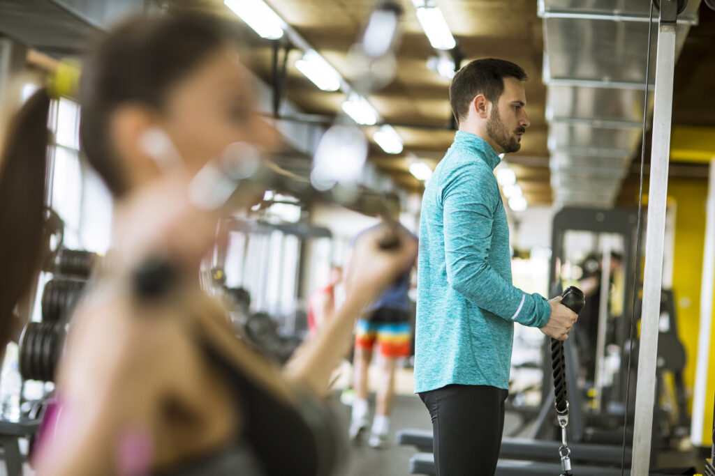 Focused man in teal athletic wear using a cable machine in a modern gym with natural lighting and other members working out nearby.