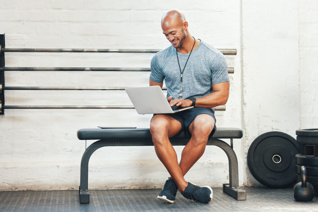 Gym owner using a laptop while sitting on a bench in a fitness studio, surrounded by weights.