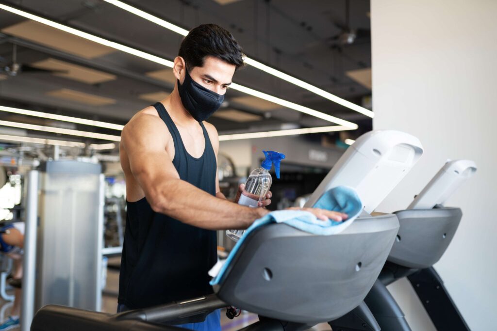 Man wearing a face mask cleaning a treadmill console with disinfectant spray and cloth.